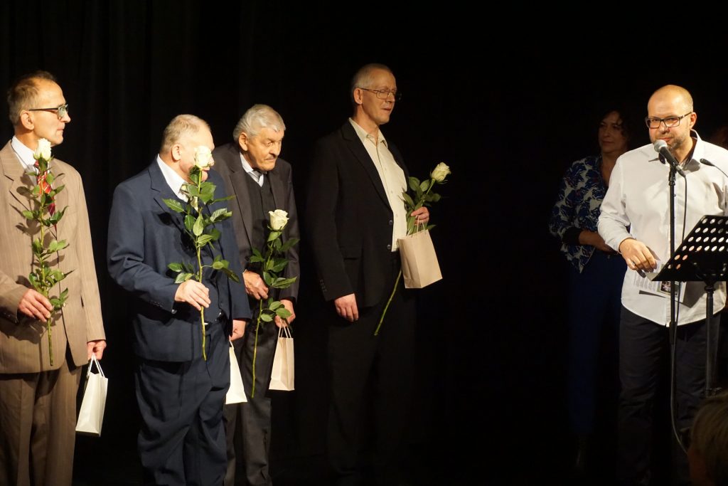 Czterech artystów trzymających róże. Przy mikrofonie prowadzący zapowiada artystów.