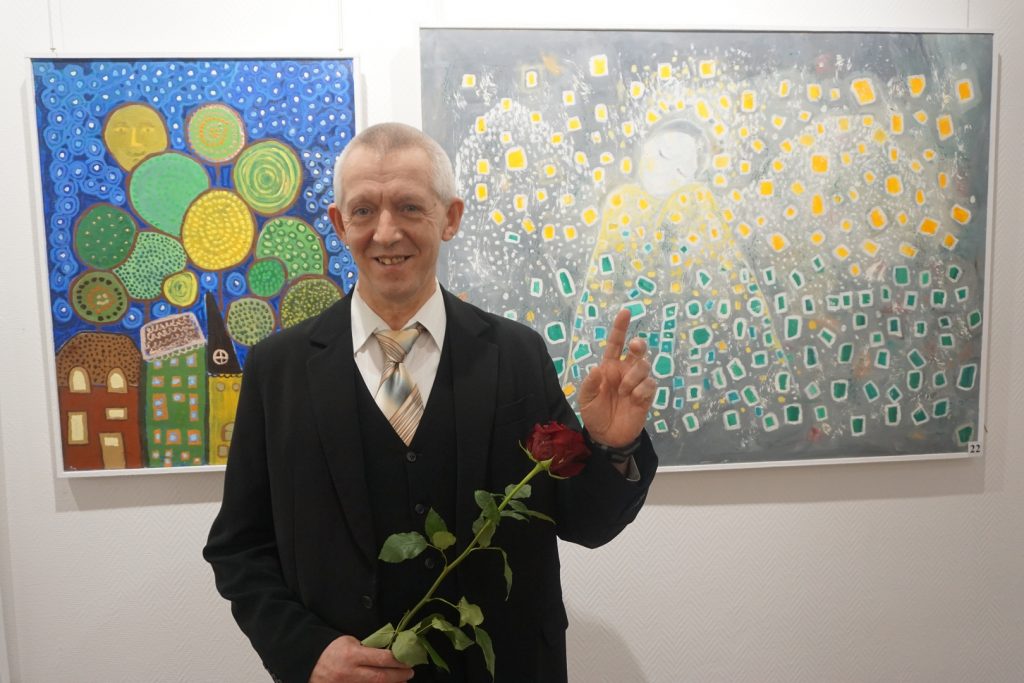 Autor obrazów a tle dwa kolorowe obrazy. W ręku trzyma róże.