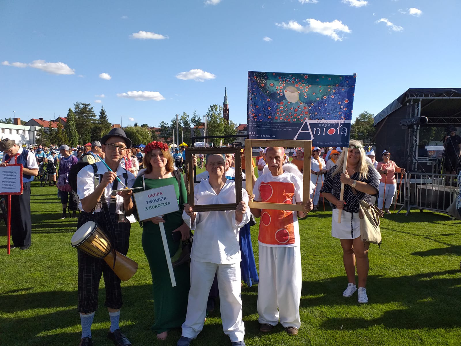 Grupa osób ubrana w Anioły promuje nadchodzący wernisaż, w tle kobiety trzymające transparent, z przodu podopieczni trzymający ramki od obrazów
