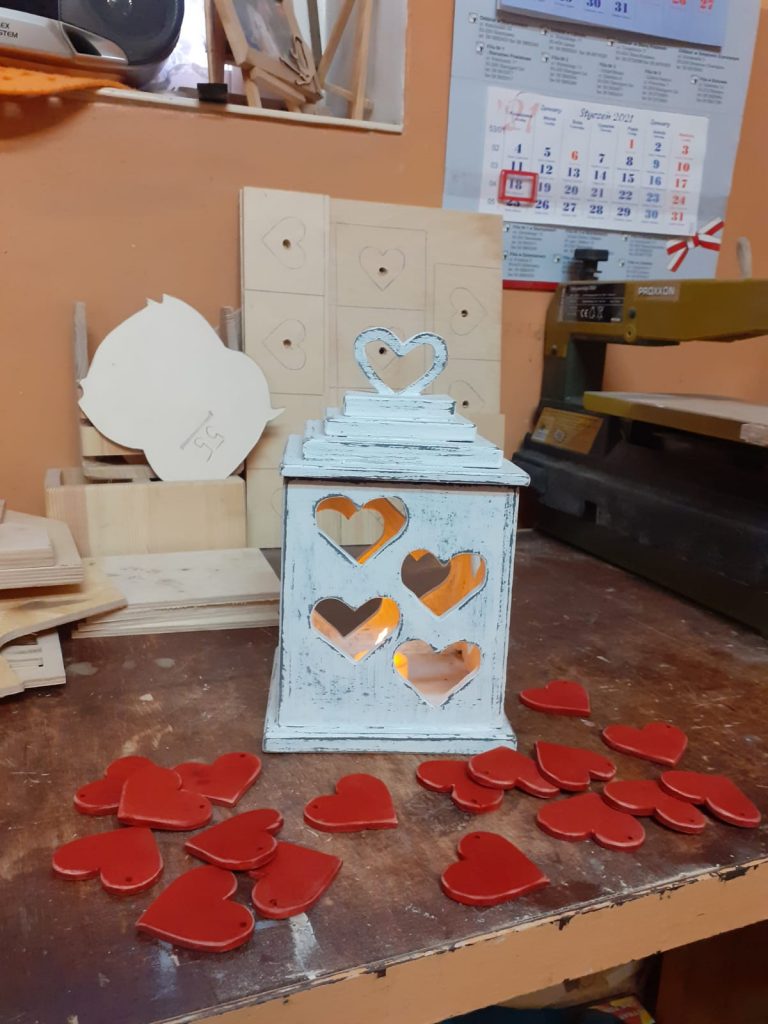 Drewniana latarnia na świeczkę z wyciętymi otworami w kształcie serca pomalowana na biało. Przy latarni leżąc drewniane, czerwone serca koloru czerwonego