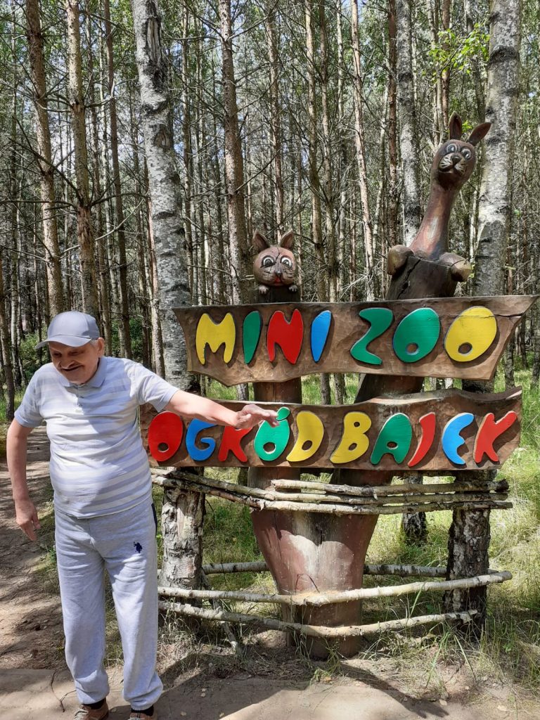 Mężczyzna stojący przy drewnianym napisie Mini Zoo oraz Ogród Bajek pokazuje ręką na tablicę