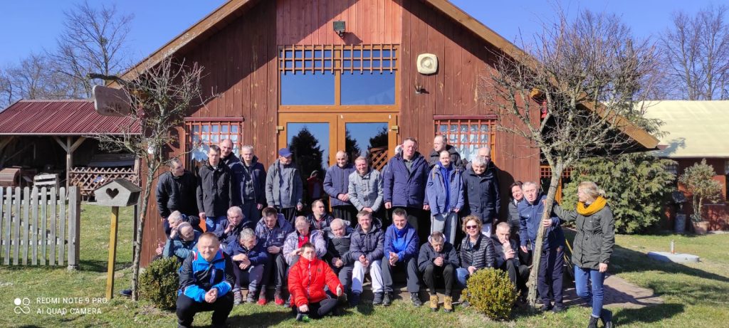 Wspólne ujęcie grupy wycieczkowej przed wysokim drewnianym budynku - Karczmą.