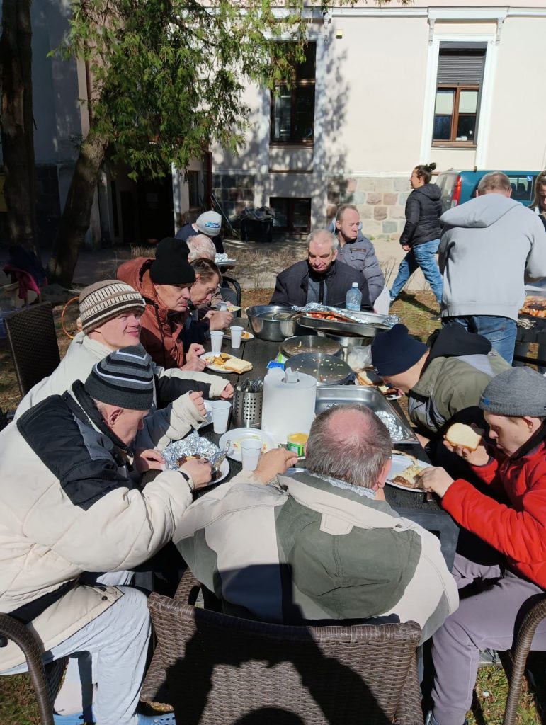 Mieszkańcy siedza przy stole na nim talerze z jedzeniem, chleb, surówki oraz mięso. Mieszkańcy ubrani w czapki i kurki. świeci słońce