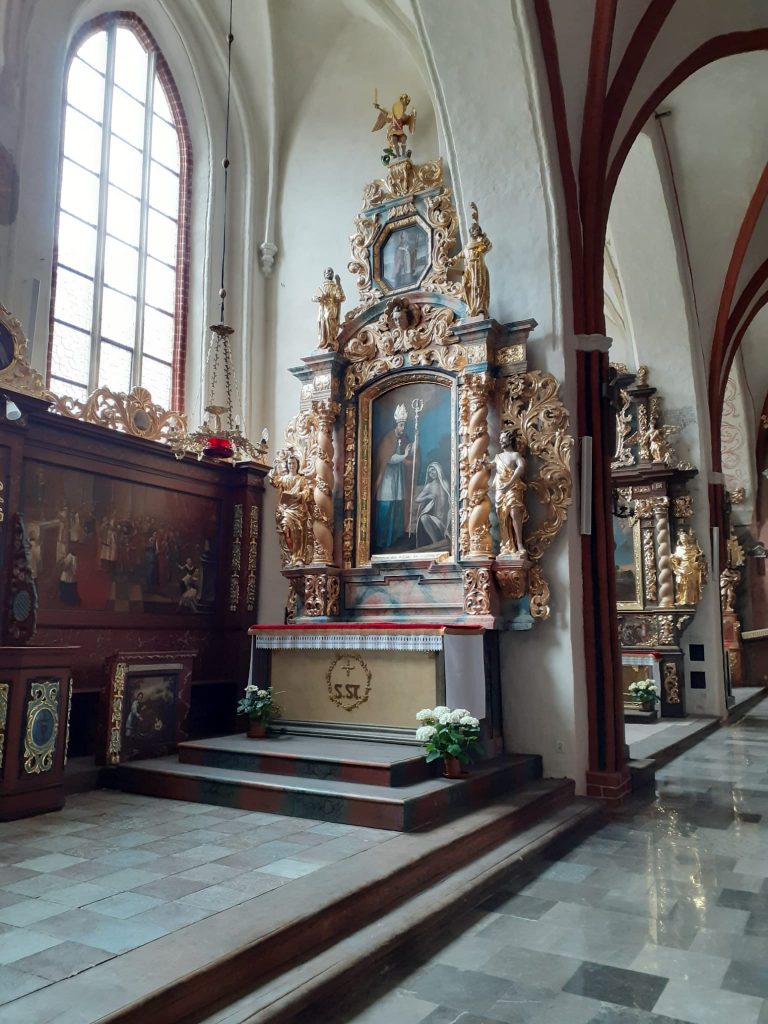 Drewniane ławki w kościele, a przed nimi obrazy ze złotymi zdobieniami.