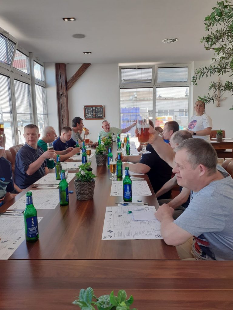 Grupa osób spożywająca bezalkoholowe piwo. Wszyscy siedzą przy drewanianym dużym stole.