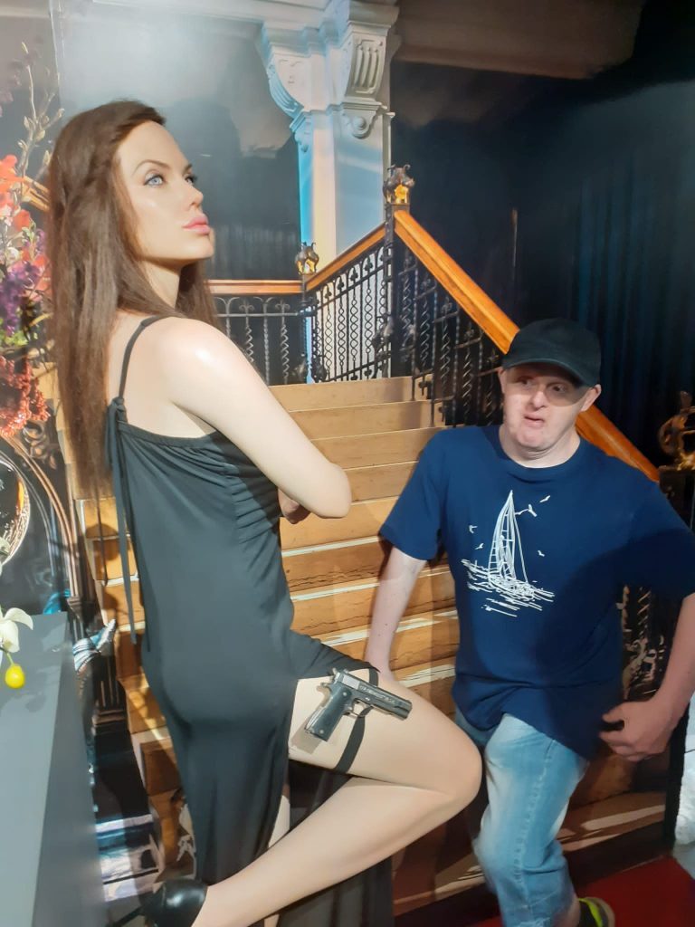 Mieszkaniec spogląda na opierającą się figurę woskową Angelina Jolie, która opiera się z założonymi rękoma, ubrana w sukienkę, przy nodze ma pistolet.