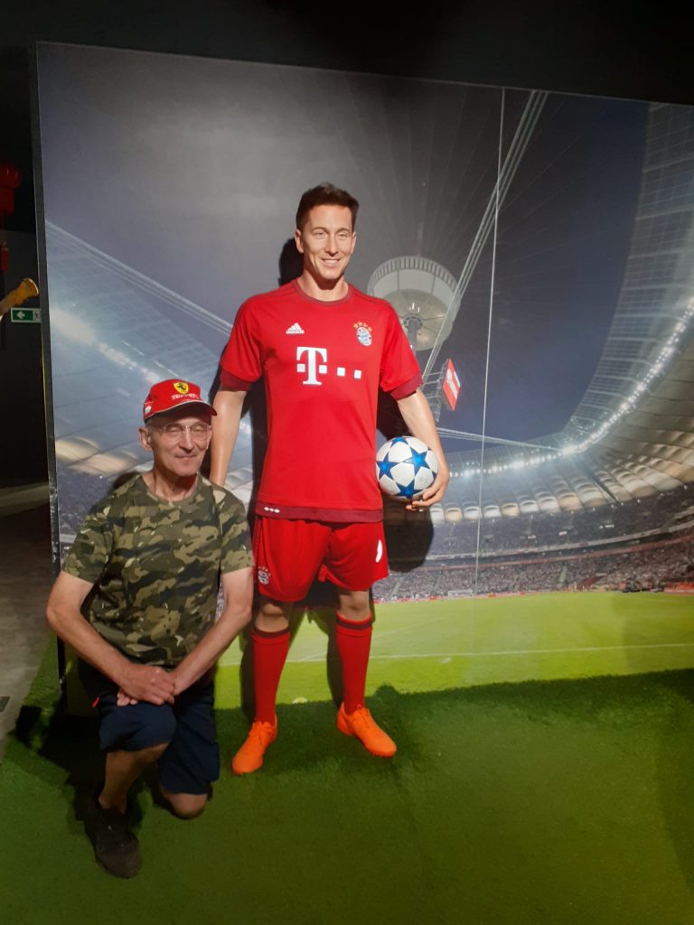Podopieczny kuca przy figurze woskowiej Roberta Lewandowskiego, który trzyma piłkę z Ligi mistrzów. Robert ubrany jest w koszulkę Bayernu.