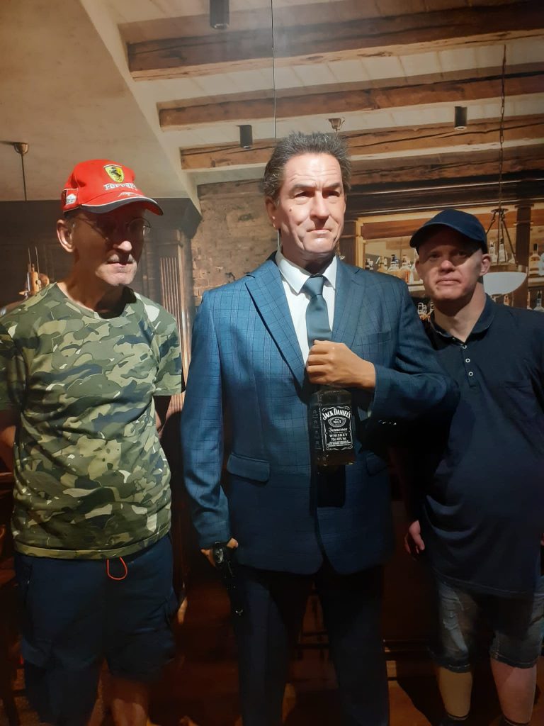 Dwóch podopiecznych stojących obok figury woskowej Roberta De Niro, która trzyma Whiskey w ręce.