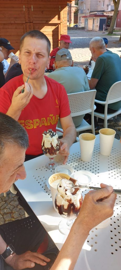 Mężczyzna siedzi przy stole i obluje łyżeczkę, obok niego kolejny mieszkaniec zajadający lody. Za nimi grupa osób jedząca lody.