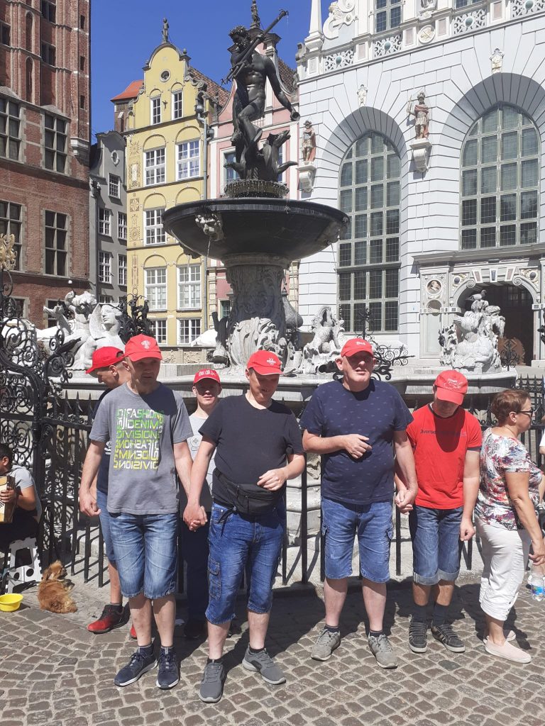 Sześciu mieszkańców opierających się o barierkę, za nimi fontanna Neptuna oraz kamienice w Gdańsku.