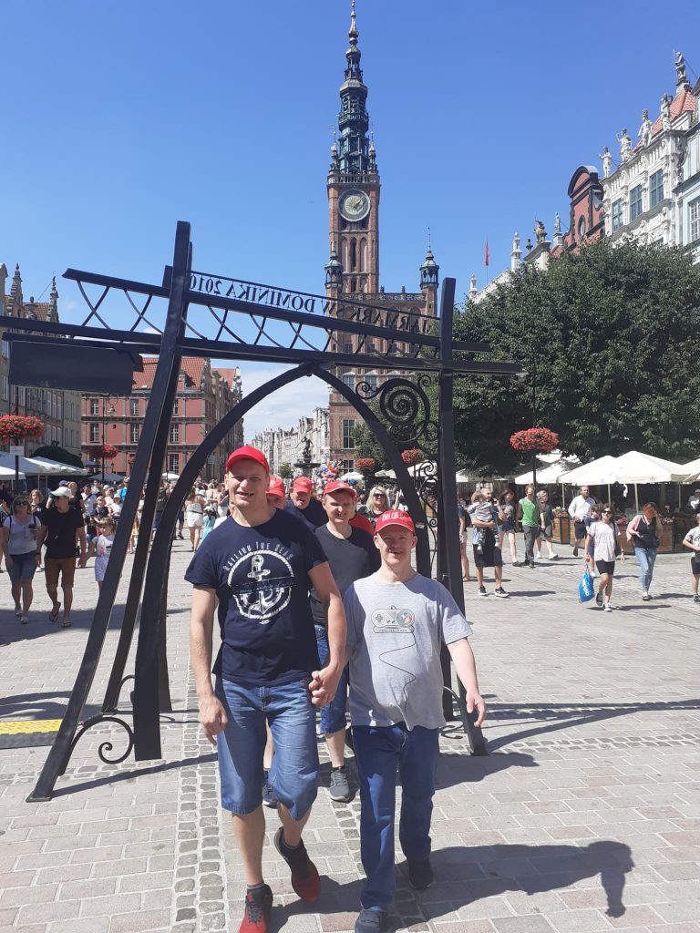 Grupa mężczyzn idąca wspólnie jednen za drugim w parach. Przechodzi przez postawioną metalową bramę na ul. Długiej. W tle inne osoby zwiedzające Gdańsk, kamienice oraz kościół.