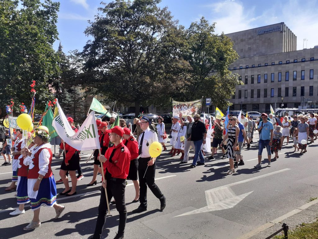 Grupa osób idąca ulicą. Niektóre z tych osób trzymają flagi, transparenty lub balony. Idzie Kociewski korowód.
