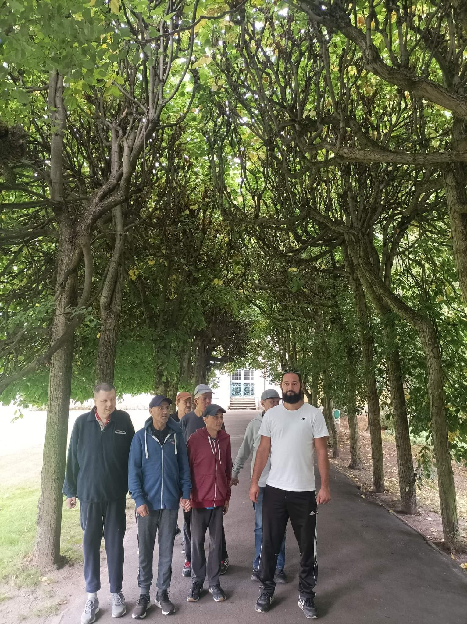Grupa mężczyzn wraz z opiekunem podczas spaceru w Parku Oliwskim. Stoją między wysokimi koronami drzew.
