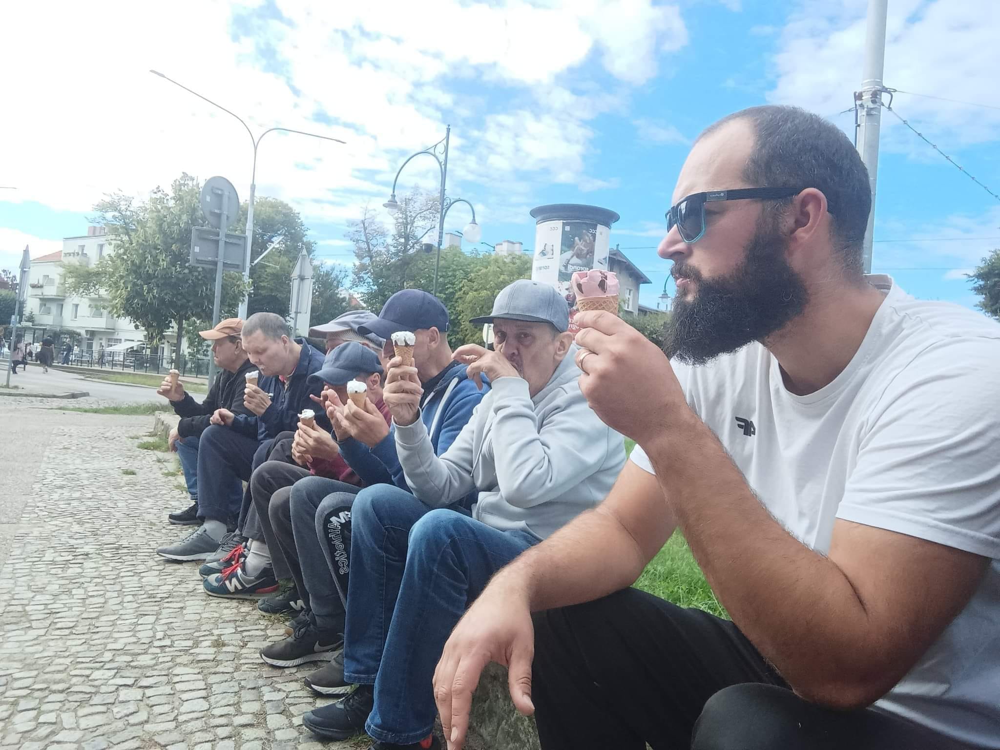 Grupa mężczyzn siedząca razem na świeżym powietrzu i jedząca lody. W tle miasto.