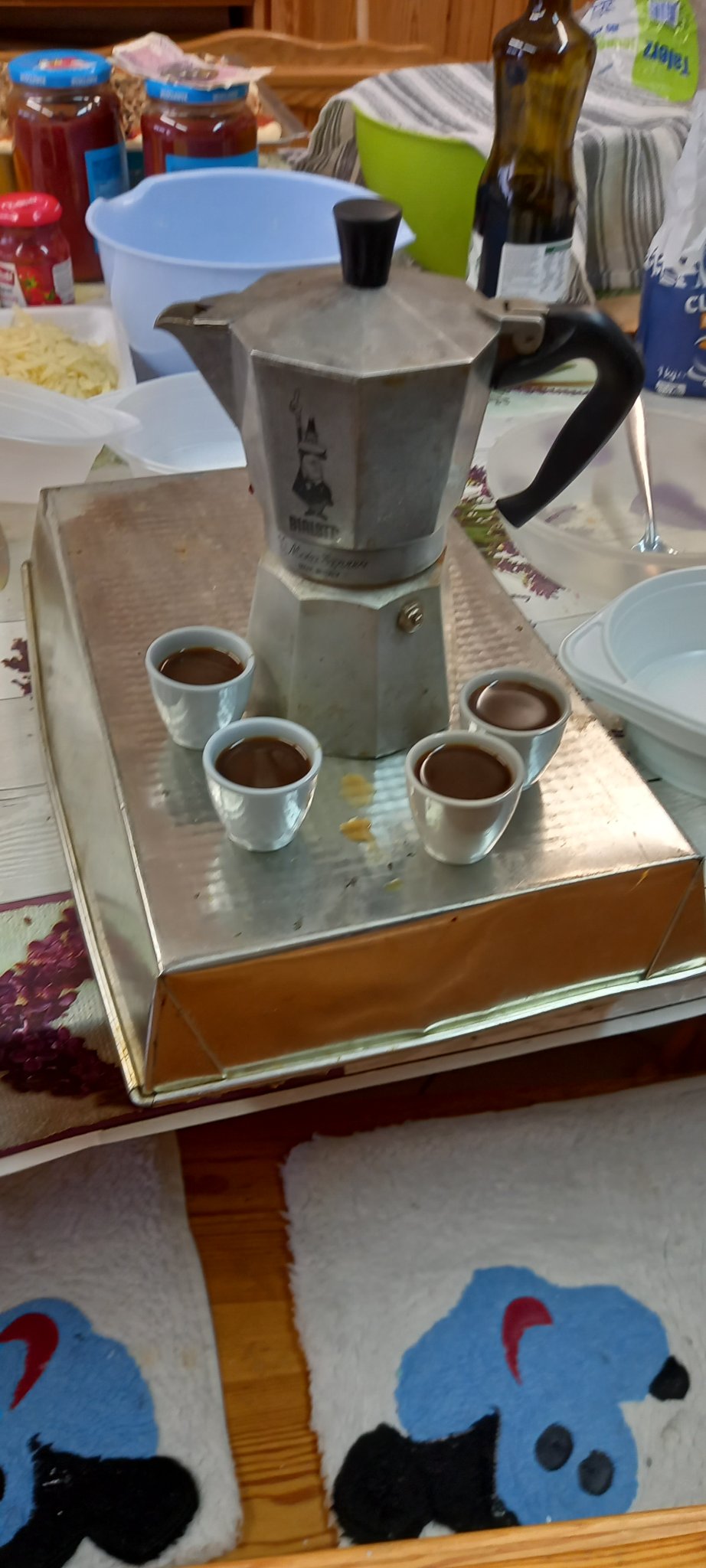 Metalowa kawiarrka ustawiona na metalowej blaszce do piczenia, obok kawiarki cztery filiżanki z kawą.