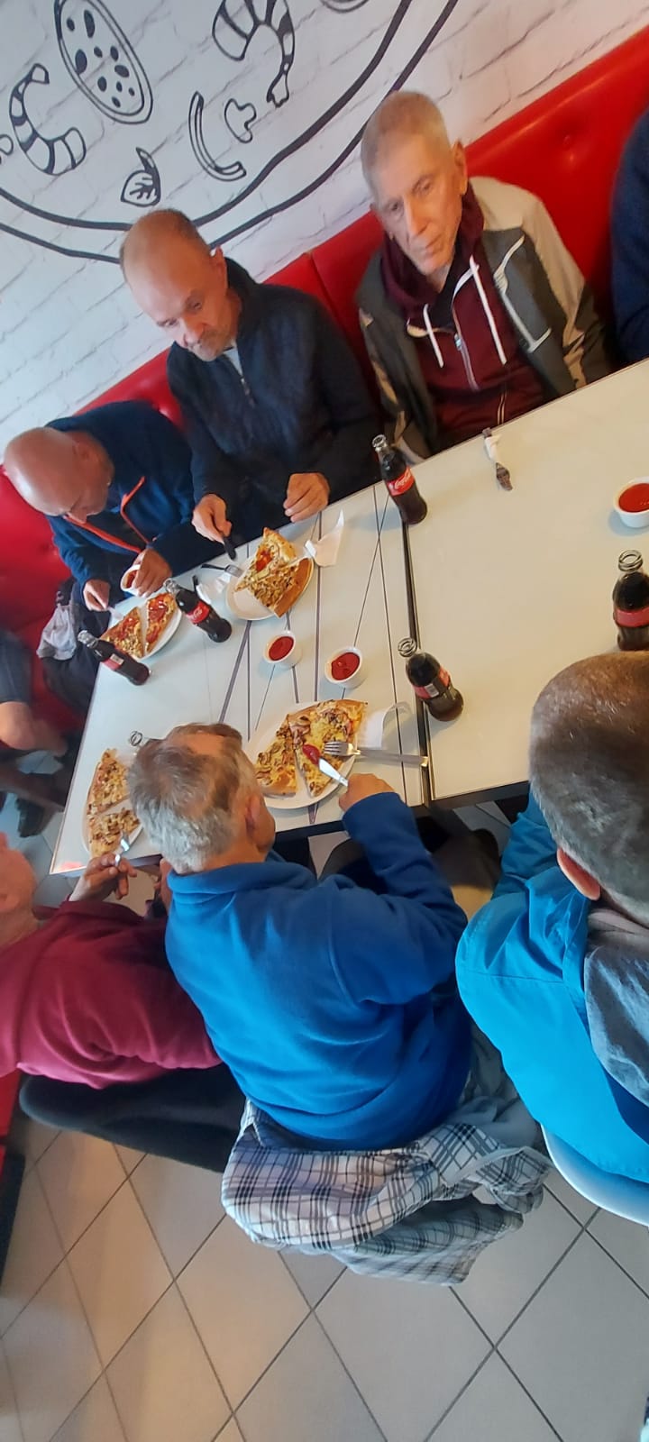 Mieszkańcy siedzą wspólnie przy stole, jedzą pizzę oraz piją coca colę.