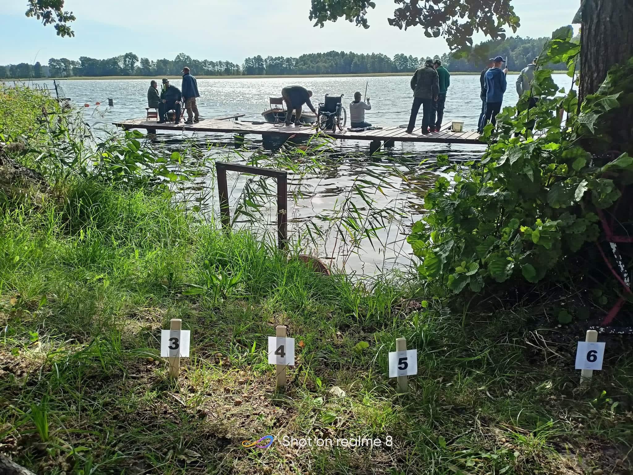 Dziesięć osób przebywających na pomoście, przed nimi jezioro i odbijające się słońce. Jest jasno. Za nimi woda oraz zielona trawa i powbijane tabliczki z numerami startujących 3,4,5,6.