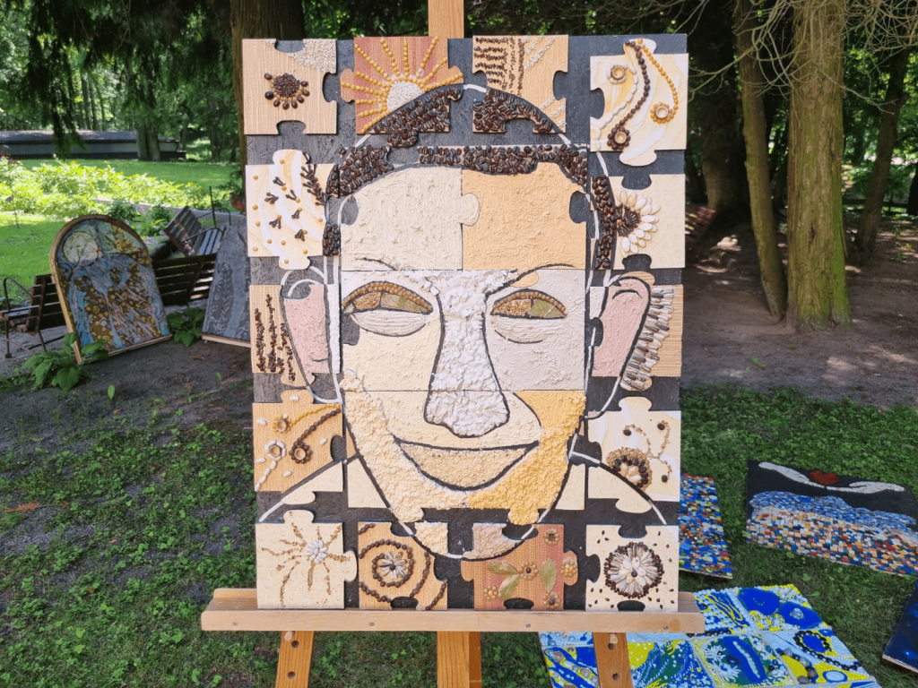 Obraz stojący na sztaludze, przedstawia uśmiechniętą twarz mężczyzny. Obraz wykonany techniką własną, płyta wiórowa, puzzle ze sklejki, pestki, ziarna, masa strukturalna, drobne kamyczki