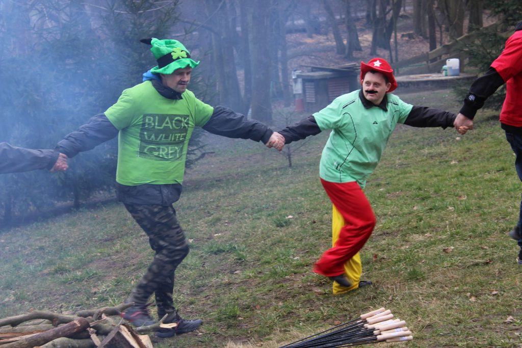 Dwóch mężczyzn trzymających się za ręce podczas wspólnego tańca przy ognisku. Podopieczni ubrani w kolorowe stroje witają wiosnę