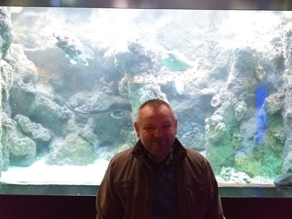 Podopieczny na tle wielkiego akwarium z kolorowymi rybami pozuje do zdjęcia. 