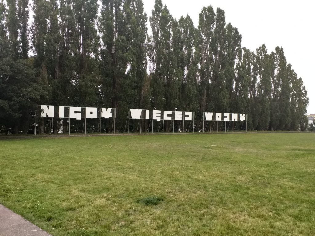 Zielona trawa, w oddali napis "Nigdy więcej wojny" za napisem wysokie drzewa.