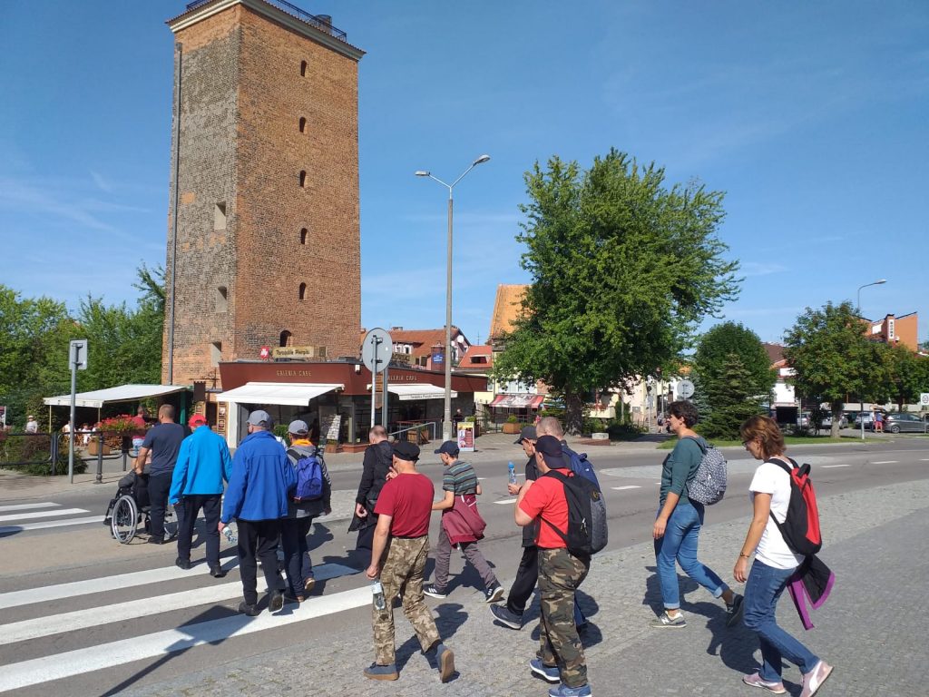 Grupa mieszkańców przechodząca przez pasy w tle wysoka czerwona wieża z wieloma oknami