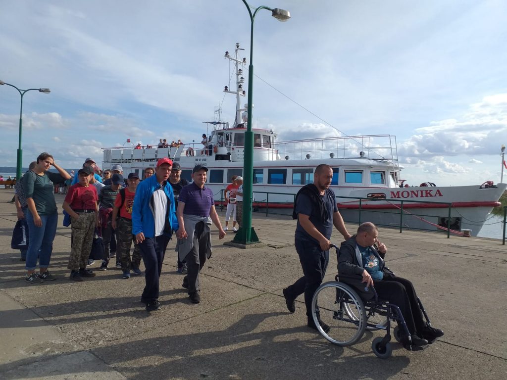 Grupa mieszkańców DPS Rokocin z przodu grupę prowadzi opiekun pchający mieszkańca na wózku