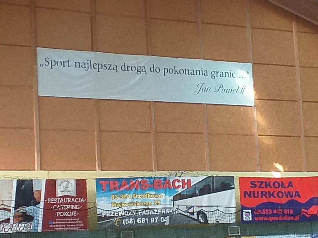 Wielki baner na ścianie hali sportowej (Cytat Jana Pawła II) "Sport najlepszą drogą do pokonania granic..." 