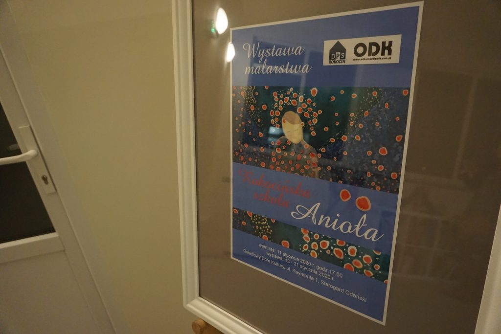 Plakat Wystawy Malarskiej Rokocińska Szkoła Anioła w białej ramce wisi na sztaludze.