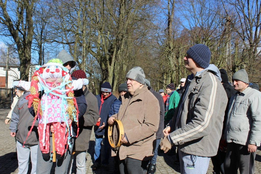 Grupa mieszkańców, ubrana w kurtki i czapki, jeden z nich trzyma tamburyno a drugi marzannę. W tle drzewa