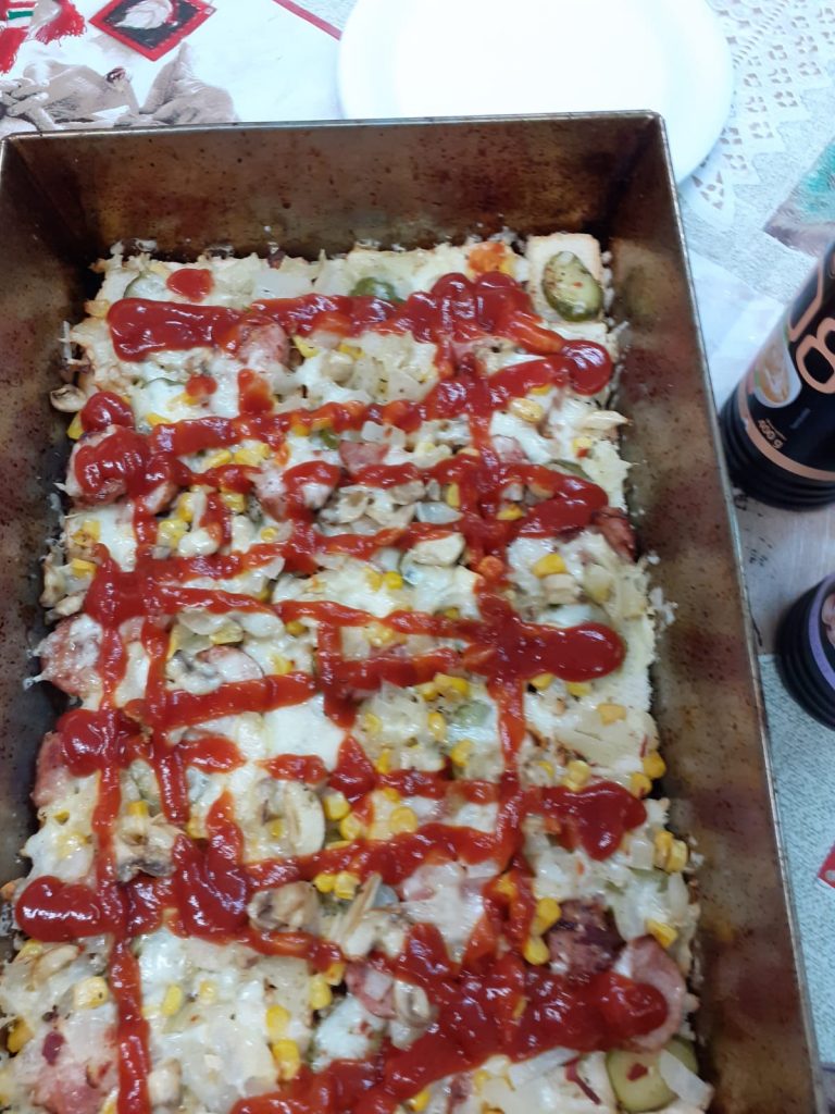 Metalowa blacha leżąca na stole, w której jest pizza polana czerwonym ketchupem