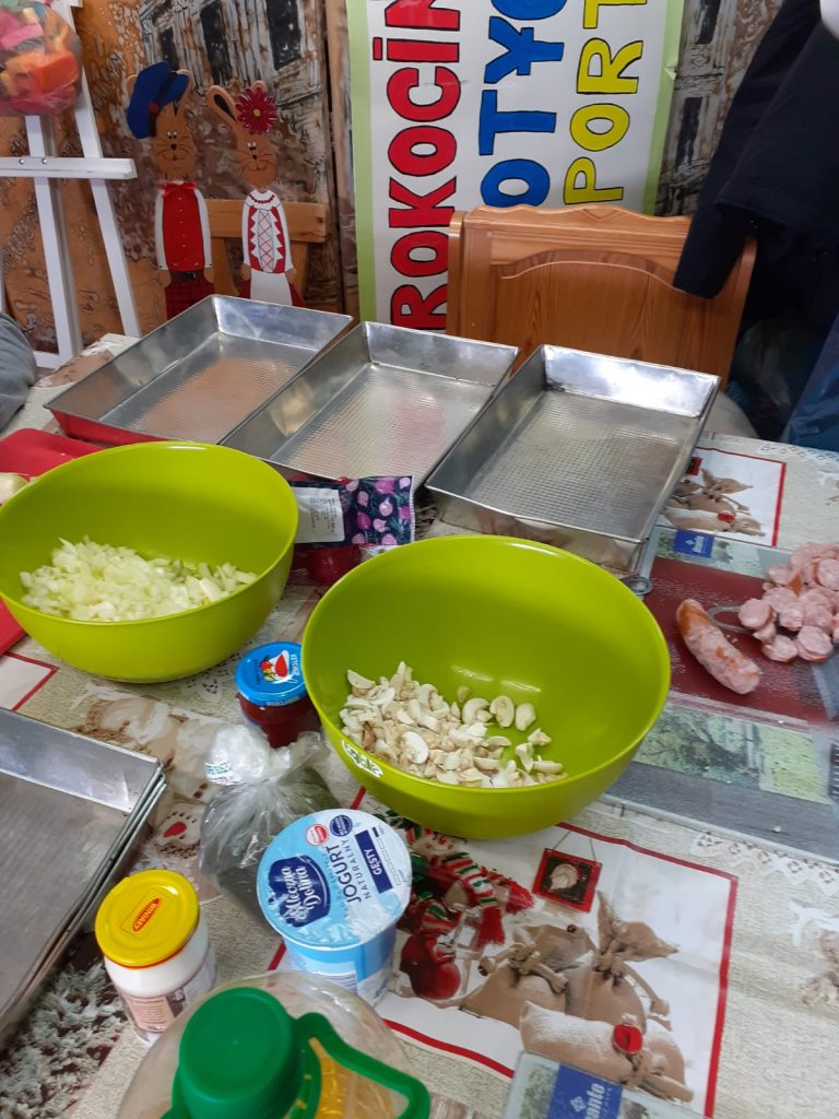 Na stole leżą trzy metalowe duże blachy, dwie zielone miski w jednej cebula w drugiej pieczarki, sosy i jogurt