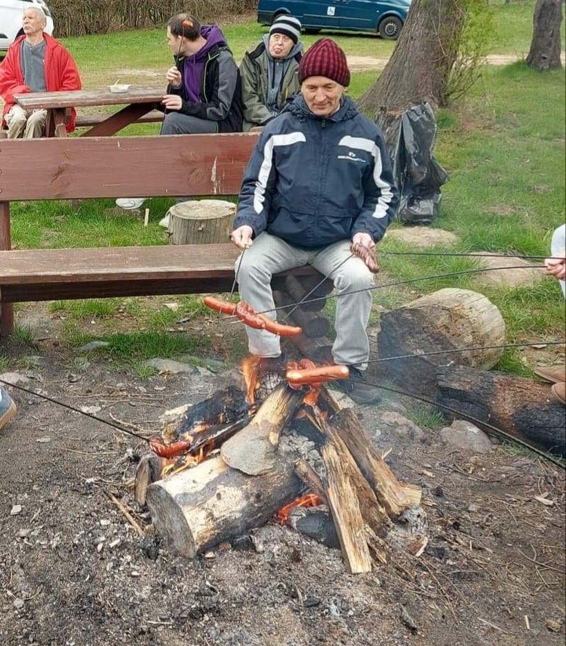 Mieszkaniec siedzi przy ognisku na drewnianej ławce w ręce trzyma kij z nabitą kiełbaską