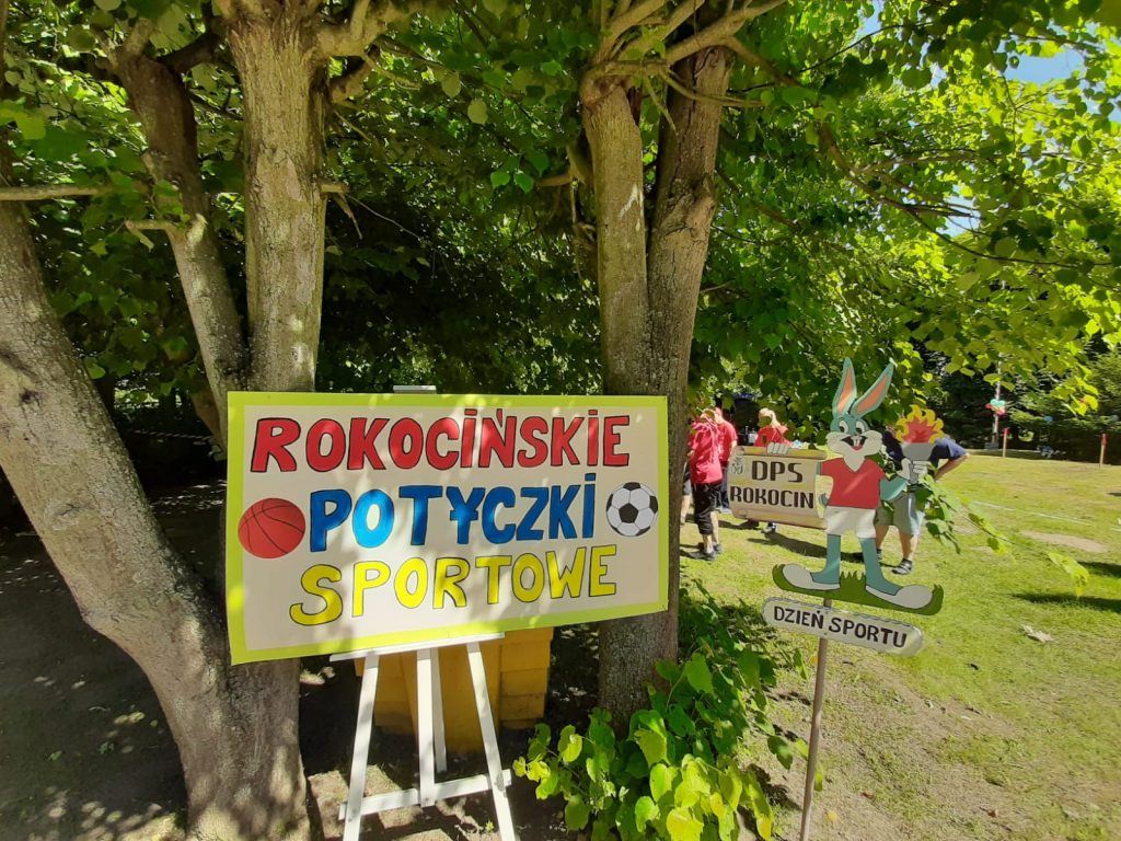 Kolorowy napis na tablicy Rokocińskie potyczki, tablica stoi na drewnianym stelarzu.