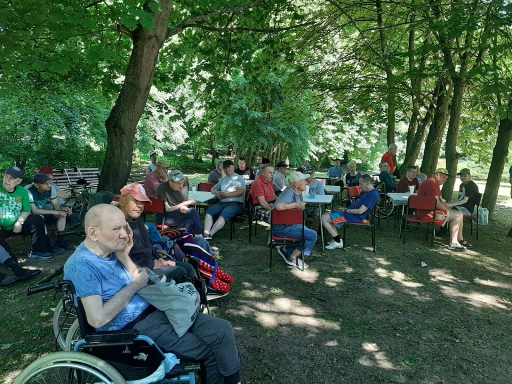 Grupa widzów siedzących na krzesłach w cieniu oraz wózkach inwalidzkich