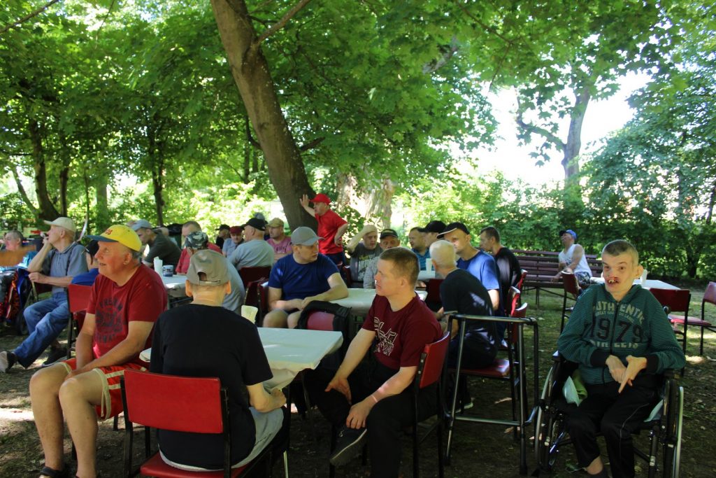 Wiele osób siedzących na krzesłach w cieniu między zielonymi drzewami