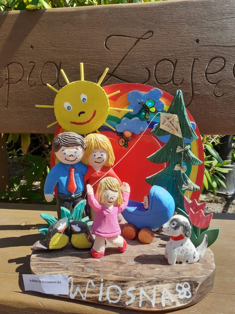 Rzeźba z drewna, przedstawia kolorowe postaci mężczyzny, kobiety, dziecka córeczki oraz dziecka w wózku obok pies a za nimi drzewo, słoneczko oraz tęcza. 