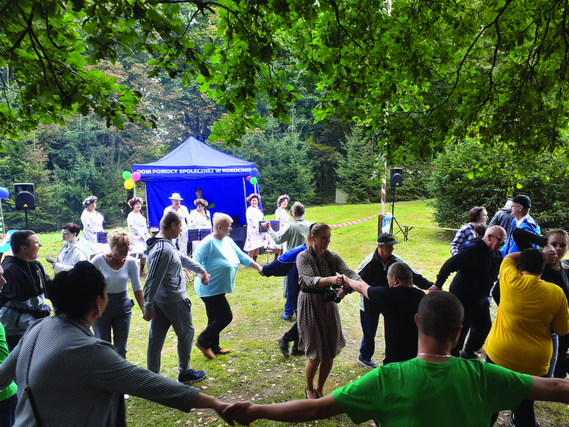 Grupa osób tańczy razem trzymając się za ręce w tle zespół folkowy