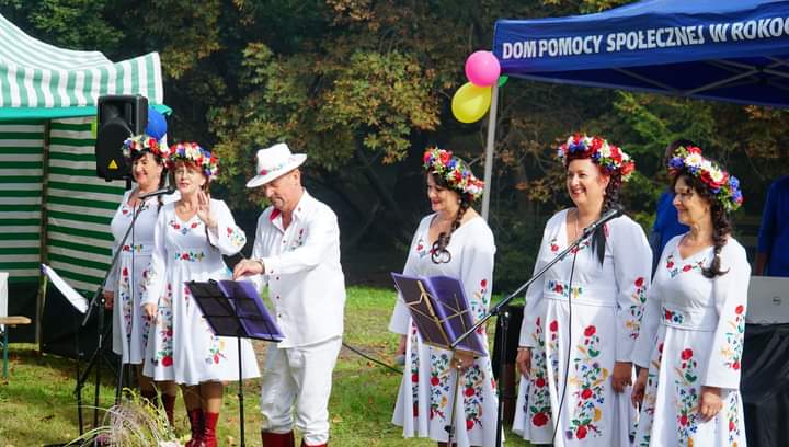 Pięć kobiet ubranych w folkowe stroje oraz mężczyzna z białym kapeluszem stoją przed śpiewnikami i mikrofonami. Razem śpiewają