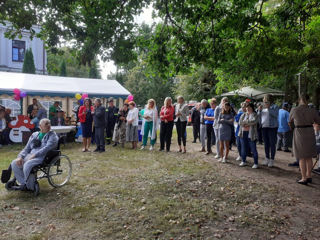 Grupa osób stojąca obok siebie na zielonej trawie. Obok duży namiot ze stołami, nad głowami zielone liście