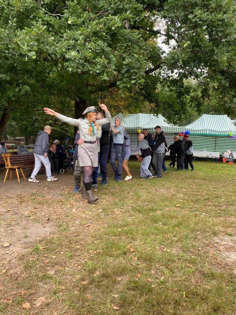 Kobieta przebrana w strój harcerza prowadzi "wężyk" w którym bawią się podopieczni z opiekunami. Idą po zielonej trawce