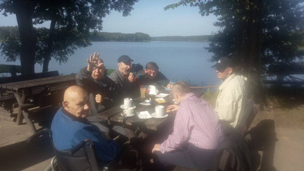 Grupa mieszkańców siedzi na drewnianej ławce niedaleko jeziora. Jedzą oraz piją kawę.