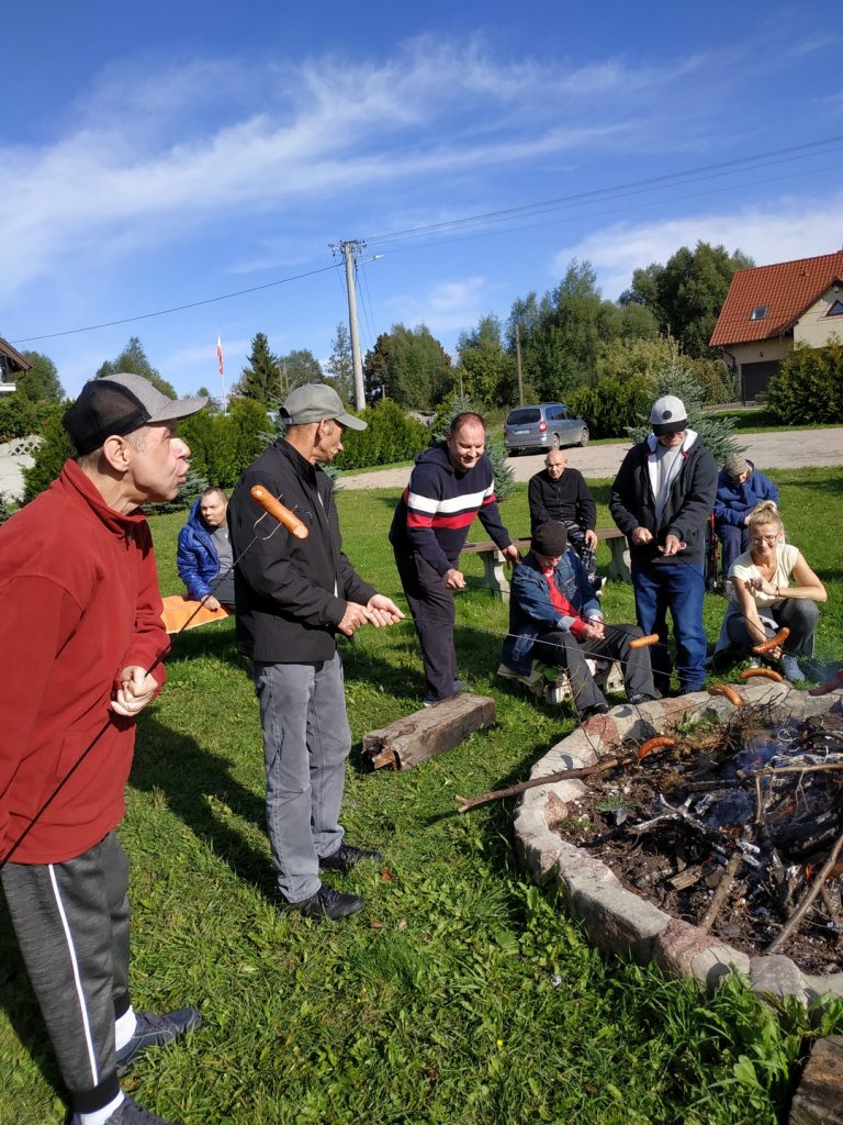 Grupa osób stoi nad rozpalonym ogniskiem, w ręce trzymają kije z kiełbaskami