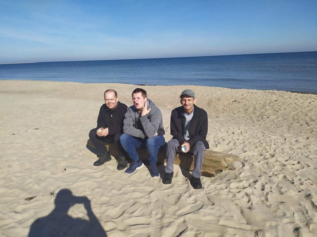 Trzech mężczyzn siedzi na drewnianej kłodzie na plaży za nimi morze Bałtyckie