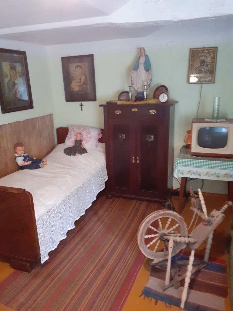 Niewielki pokój w starodawnym stylu. Ciemna komoda, obrazy dzieci oraz matki Boskiej na ścianie, łóżko z dwiema lalkami oraz stary mały telewizor na stoliku