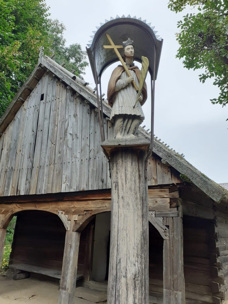 Drewniany domek ,a przed nim na drewnianym palu kapliczka z postacią trzymającą krzyż.
