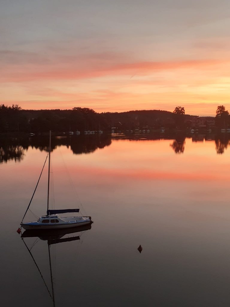 Zachód słońca nad jeziorem. Na jeziorze odbija się w tafli wody las oraz pomarańczowe niebo. Jedna łódka na wodzie.