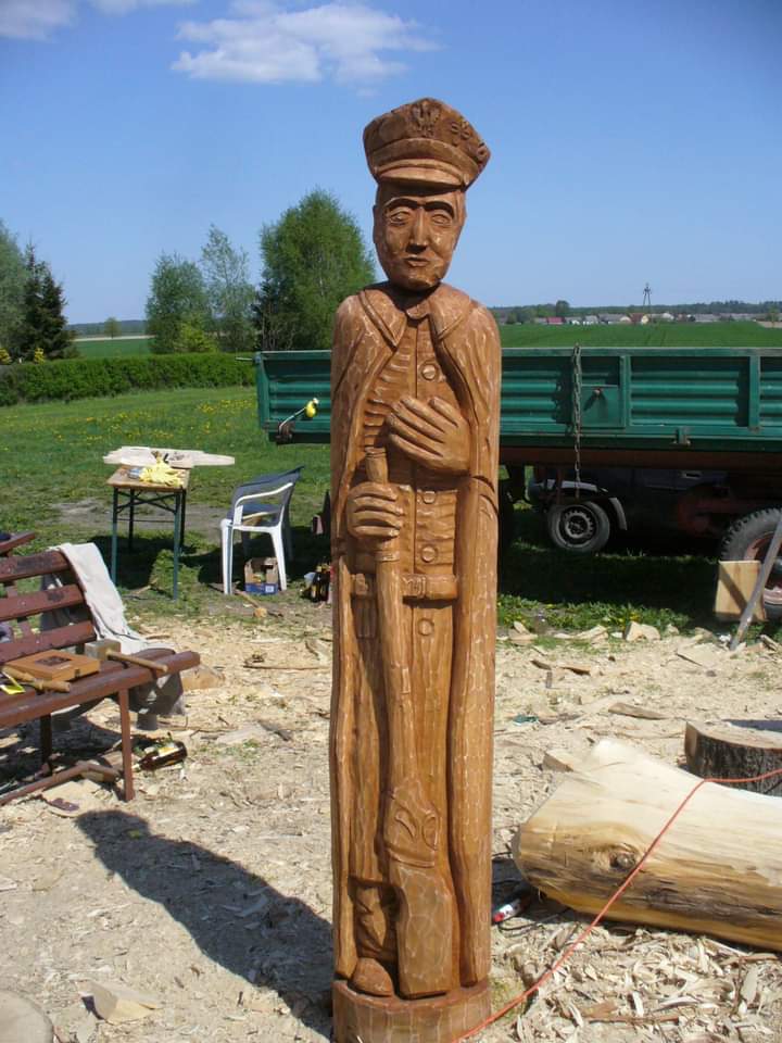 Drewniana rzeźba Powstańca Wielkopolskiego po skończonej obróbce ręcznej. Z tyłu rozrzucone drewno, ławka i samochód