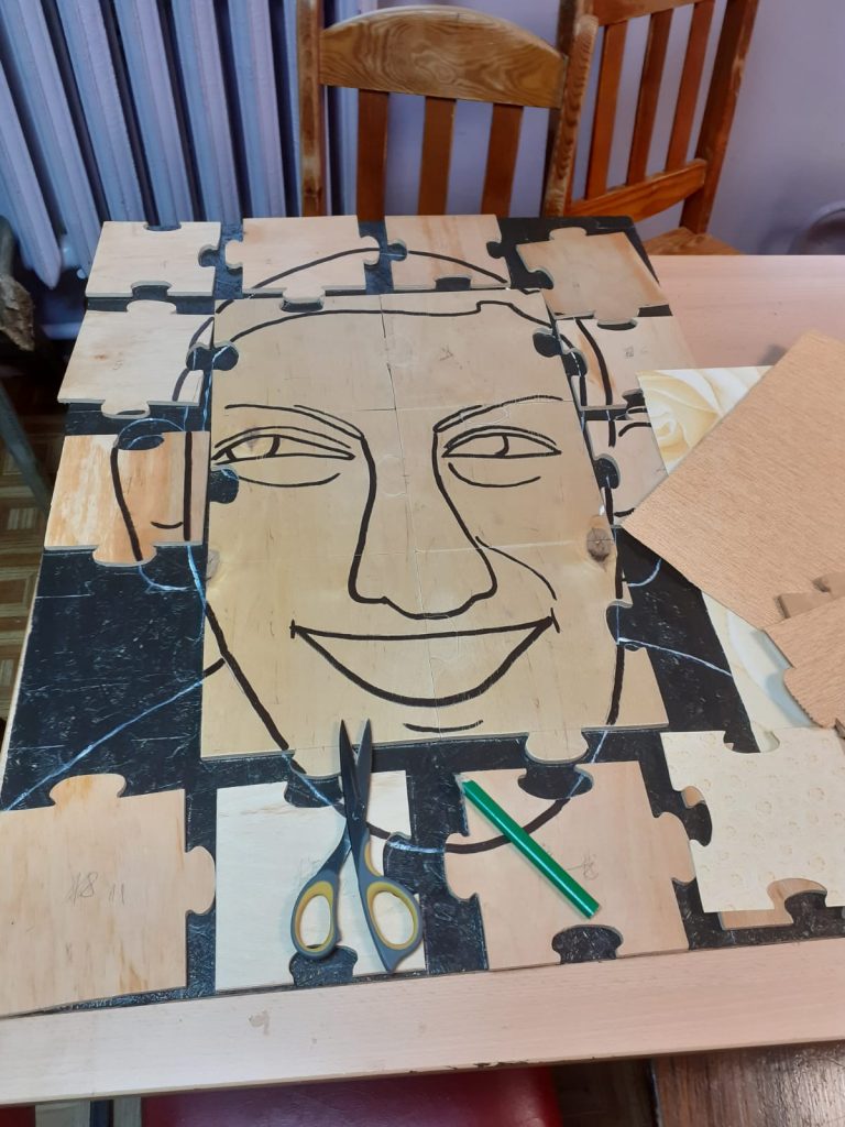 Duże drewniane elementy, wycięte na wzór puzzli, obraz do składania, na nim namalowan uśmiechnięta twarz mężczyzny