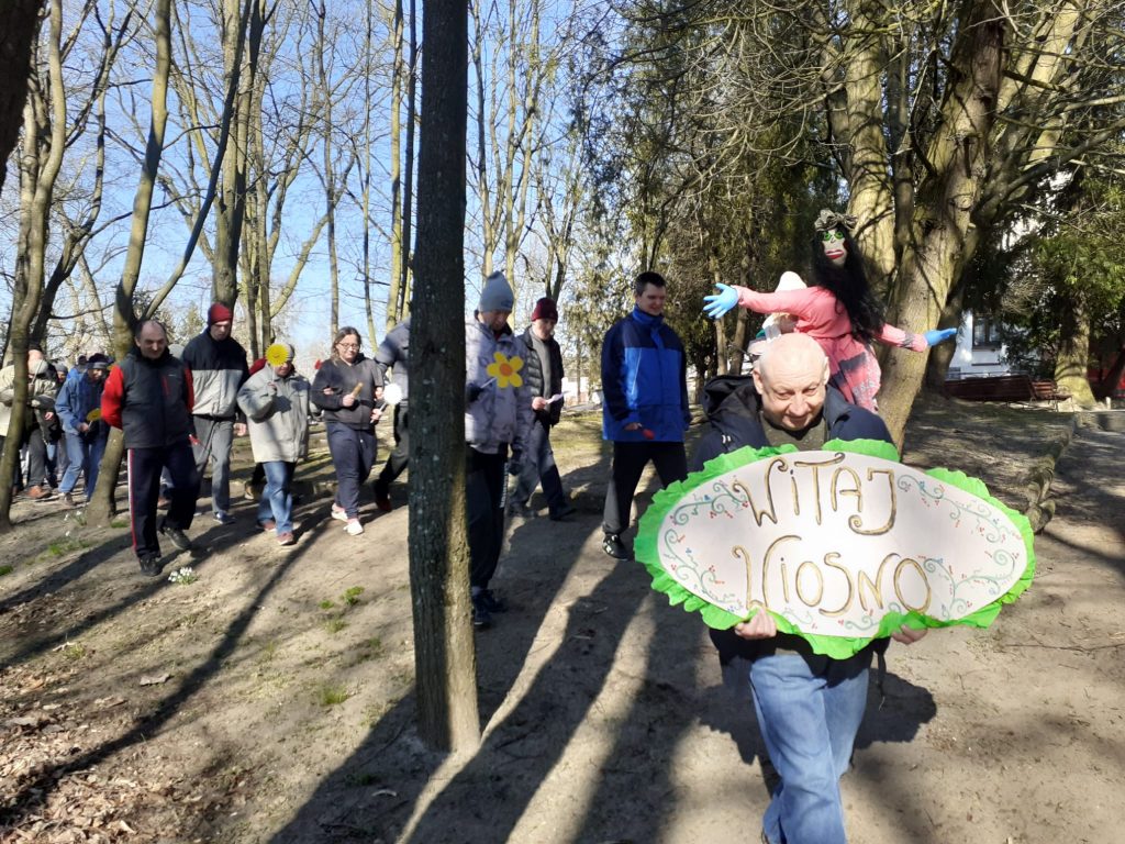 Mężczyzna idący z napisem wykonanym z drewna "Witaj wiosno" za nim idą inne osoby. W koło las oraz świeci słońce